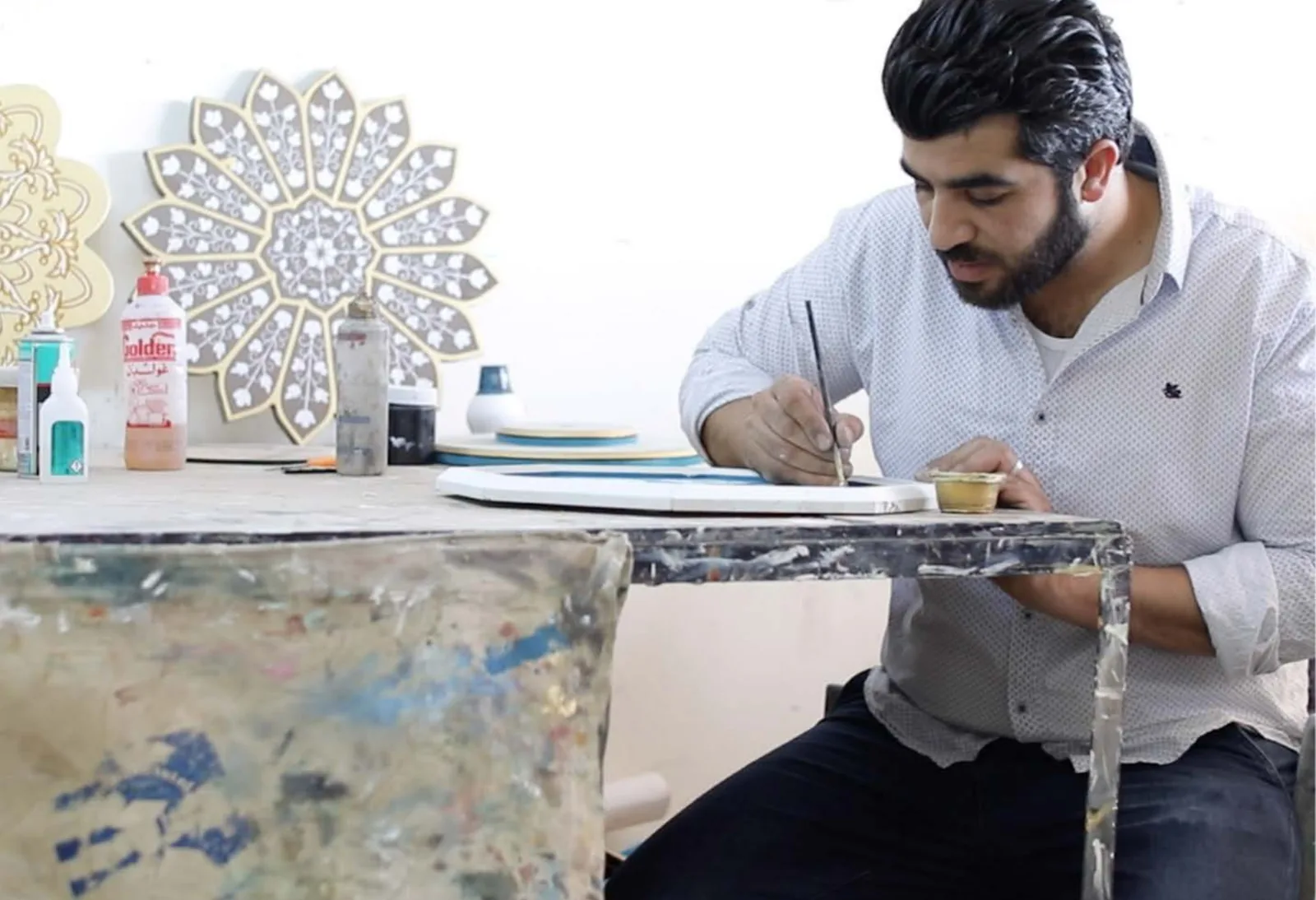 A woodworker painting the edge of a lacquered panel at a paint-spattered workbench, finished mandala pieces on the wall behind him