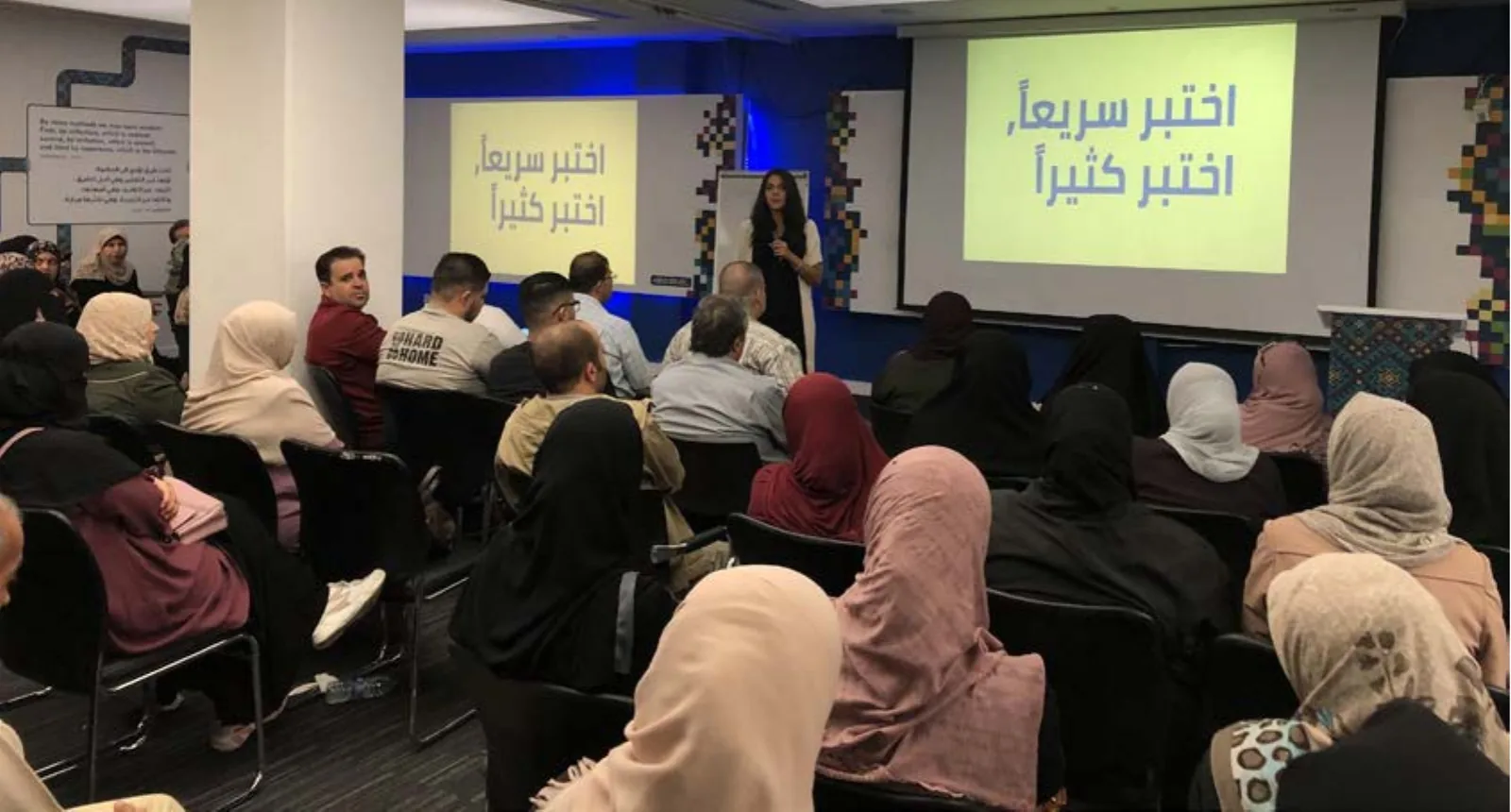 A full room of business owners, most of them women, seated facing a presenter and two screens reading test fast, test often in Arabic