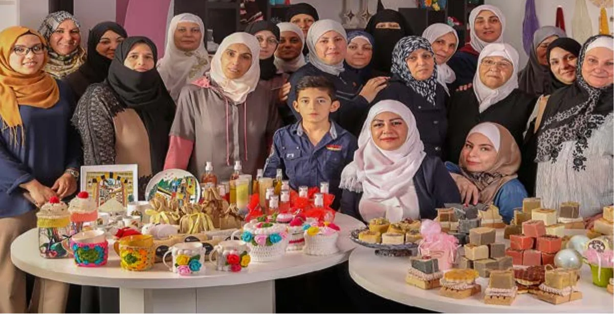 Around twenty women and one child standing behind two tables covered with handmade soap, crocheted goods and preserves