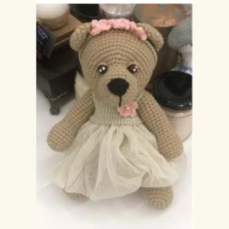 ⁨A crocheted bear in a lace dress and dust-coloured yarn, photographed on a shelf⁩